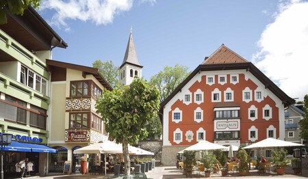 Malerischer Marktplatz in einem österreichischen Alpenort mit buntem Rathaus, Straßencafés und Kirchturm unter blauem Himmel.